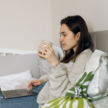 The Caucasian woman is lying on the bed and using a laptop to working online from home while her child is in another room with father