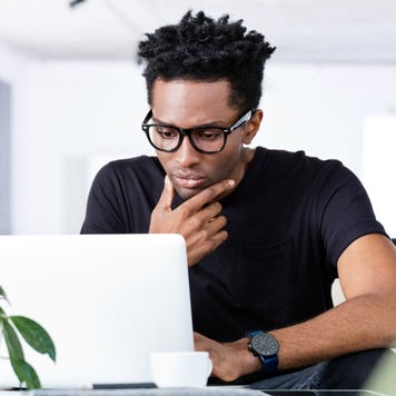 Young Black man in glasses sitting at a table and looking thoughtful while using his laptop