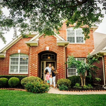 family of 4 posing in front of new red-brick house, green lawn and nice landscaping