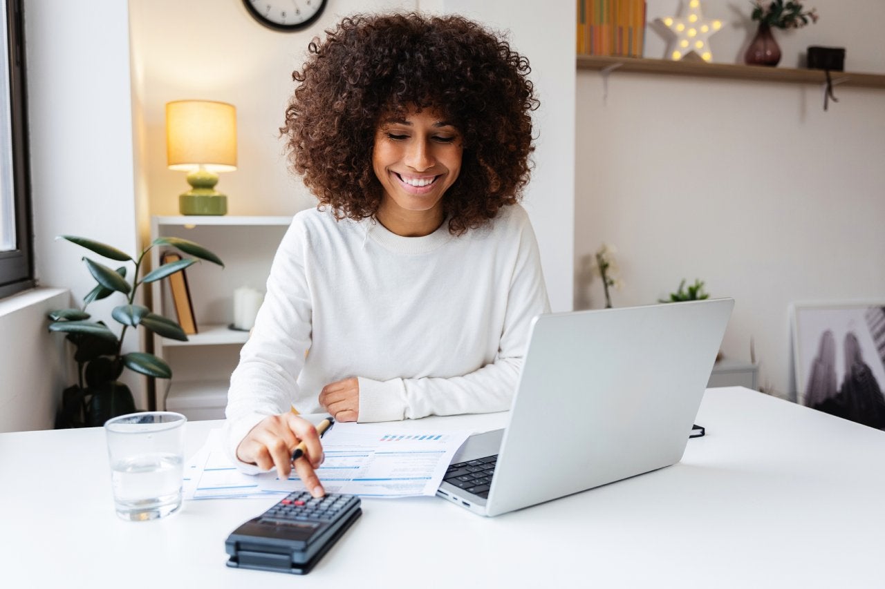 Young Latin American woman calculating expenses sitting on desk, using a calculator and laptop for her home finances