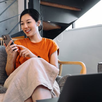 Young Asian woman using smartphone while relaxing on armchair, checking her bank account via online banking and managing her finances. Online personal banking. Wealth management. Digital banking habits. Smart banking with technology