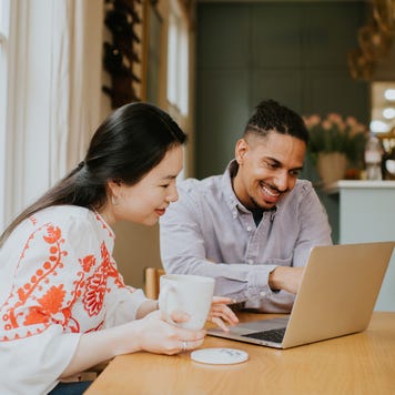 A man and a woman sit at a table. They sit behind a laptop and look relaxed and have a discussion. The man points at the computer screen.