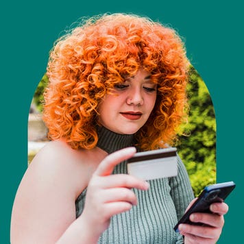 A woman with red curly hair looking down at a credit card in her right hand with a cellphone in her left hand.