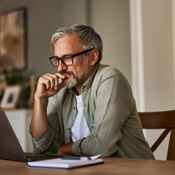 A focused mature male freelancer sitting at the wooden table at home and reading online projects on a laptop.