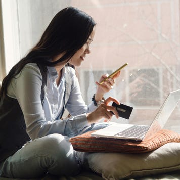 A middle-aged asian woman in blue jeans sitting on the bed in a yoga pose in front of a laptop. Remote work at home. Online shopping in quarantine period