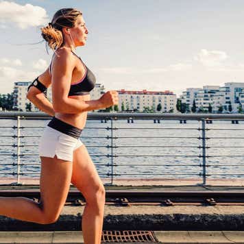 A picture of a women running and exercising beneath a clear sky, along the spree river in berlin. She is wearing sport dress and listen to music over headphones. In the distance below are some buildings and the sun is setting giving a nice warm light.