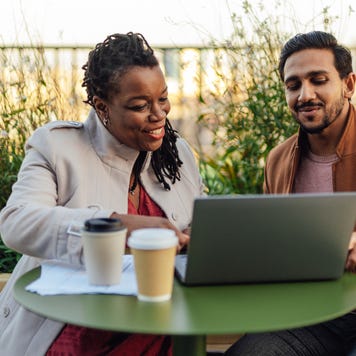 A woman and a man sit outside at a small table with a laptop and coffee in front of them. Both are smiling as they talk.