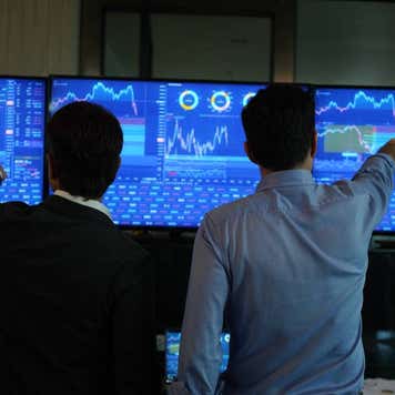 Two traders watch stocks markets on three screens