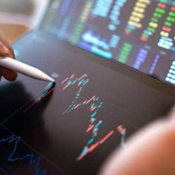 A hand holding a stylus up to a tablet displaying a stock chart.