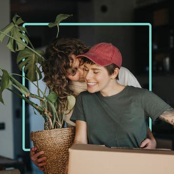 A woman is kissing her partner’s cheek while carrying a plant. Her partner is carrying a brown moving box.