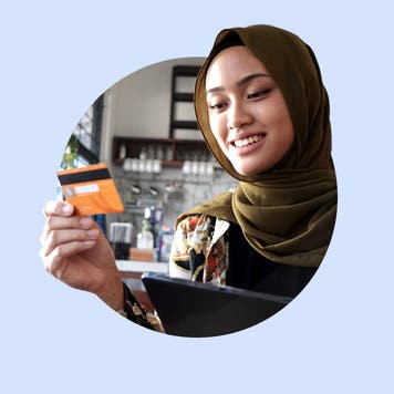 Smiling Muslim woman in a hijab looks at her credit card. Image is on top of a light blue-purple background.