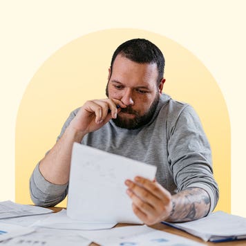 Man thoughtfully reviewing a document at a table that has many papers on it. The image has a light yellow background with a darker yellow circle behind the man.