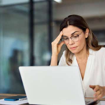 Woman with glasses in front of a computer with her hand by her head, looking worried.