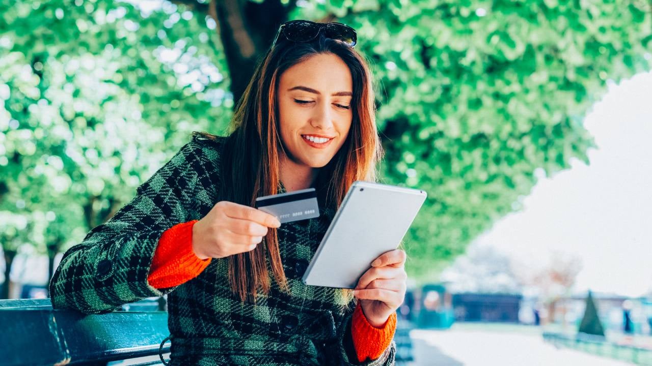 Young woman sitting at bench in park and using credit card for online shopping