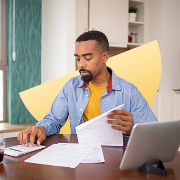 Young Black man uses a calculator at his table while looking through papers.