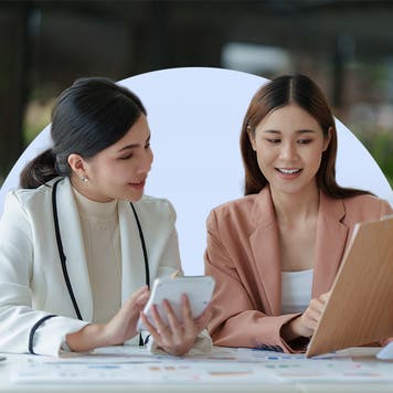 Two smiling Asian women dressed in business attire review documents on a clipboard while working in a cafe. The image includes an off-white half circle behind the two women.