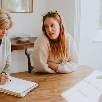Two younger woman sit around a dining table with an older woman who takes notes on a notepad.