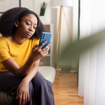 Young Black woman sitting at home looking thoughtfully at her phone.