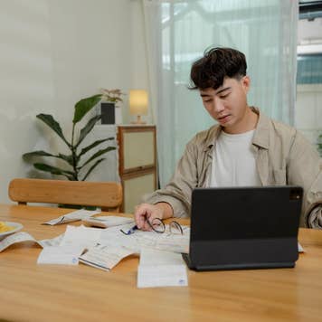 Asian male sits at dining room table with laptop and financial reports doing monthly budget to do monthly finances, pay taxes and save money for the future.