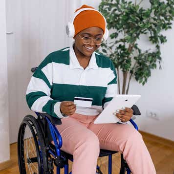 A young African American woman in a wheelchair is holding her tablet and credit card while shopping online.