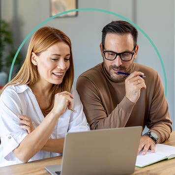 A man and a woman sit at a desk while looking at a laptop.