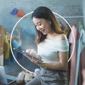 A woman, with shoulder length brown hair, leans on a desk while looking at the tablet in her hands.