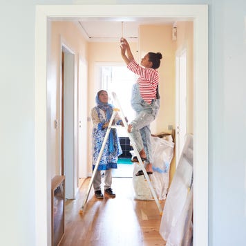 Older woman holds a ladder for a younger woman while she changes a light fixture.