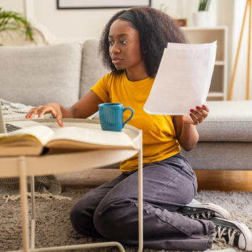 A focused woman in a yellow shirt examines a bill while working on her laptop, sitting on the floor with various documents around her, in a welcoming living room setup