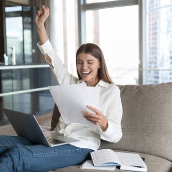 Cheerful young student woman excited with good news, reading paper document at laptop, resting on sofa at home, making winner hand, fist, smiling, celebrating success, laughing