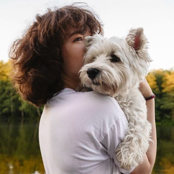 A woman stands beside a lake holding a white puppy.
