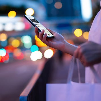 Young woman carrying a paper shopping bag, managing online banking on smartphone in city street against illuminated street lights. Tracking and planning spending. Transferring money, paying bills, checking account balances. Smart banking with technology