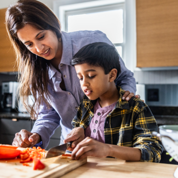 Mother helping young son cut vegetables in kitchen.