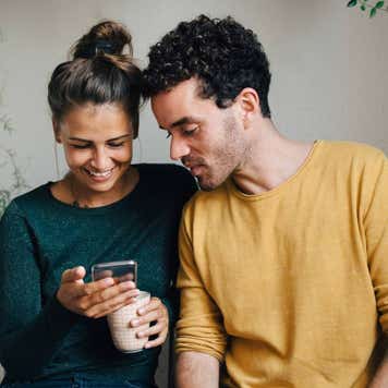 Smiling girlfriend showing smart phone to boyfriend while having coffee in living room