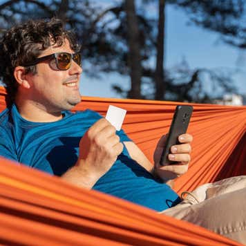A portly white male wearing sunglasses is relaxing on his hammock outside, smiling in the sun holding a debit card in one hand and a smart phone in the other.
