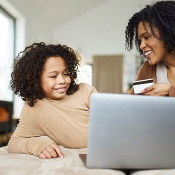 Happy African American single mother and daughter using credit card while shopping online over laptop at home.