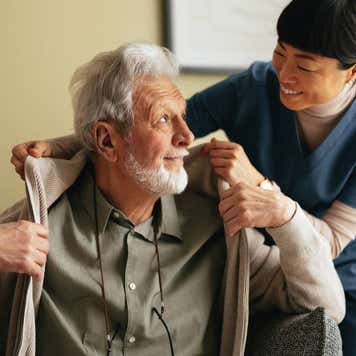 Close up shot of a smiling elderly man sitting on the sofa and a his caregiver. She is smiling while helping him put on or take off his jacket.