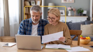 Image of a smiling older man and woman sitting close together at a table looking at an open laptop. There are papers and an open notebook on the table.