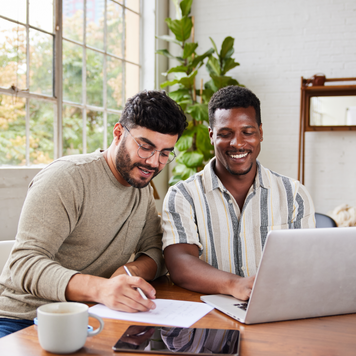 Young multiracial gay couple smiling while going over their home finances together at a table in their living room at home.