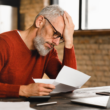 Sad, depressed caucasian man holding documents, sitting at computer in home office.