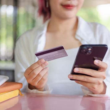 A cropped image of a young woman who sits at a table in a cafeteria holding a credit card and a smartphone