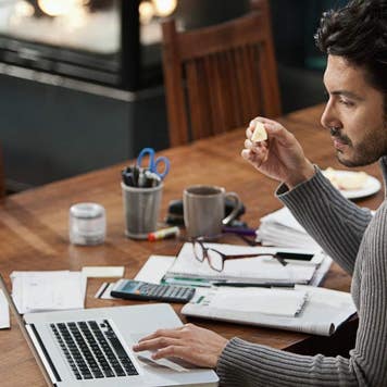 Younger man in his 30s paying bills on his computer, with calculator, papers, bills, coffee and a pair of glasses on the table by his side.