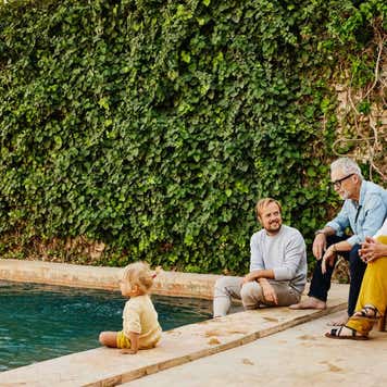 Wide shot of smiling grandparents relaxing with son and granddaughter beside pool at tropical villa during multigenerational family vacation