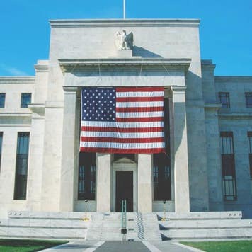 A wide-angle image of the front of the Federal Reserve HQ in Washington, DC. The U.S. flag is prominently displayed on the front of the building.