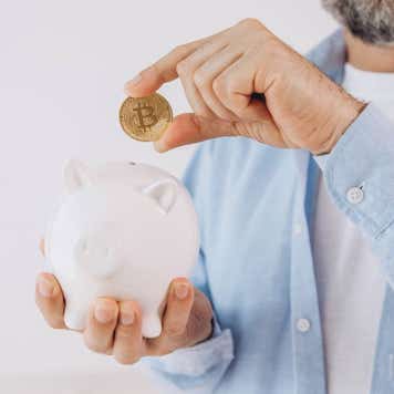 A man with a gray beard holds a bitcoin token over a piggy bank.