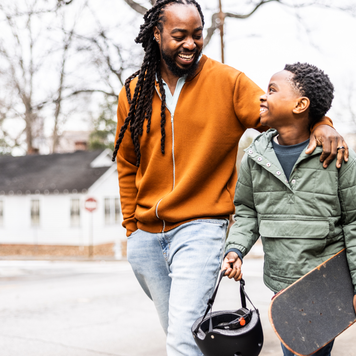 Father and young son with skateboard walking down the street.