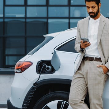 A young Black man in a suit uses his phone while charging an electric car.