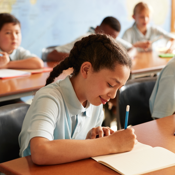 Girl with braided hair writing in book while sitting at desk with students in classroom.