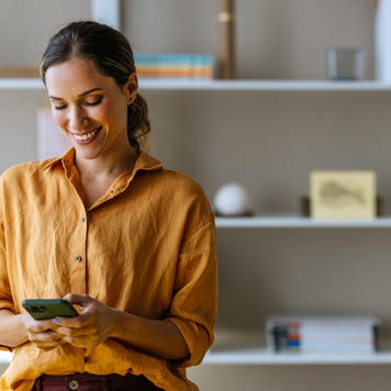 A woman looks at her investing portfolio on her smartphone in a living room with shelves behind her