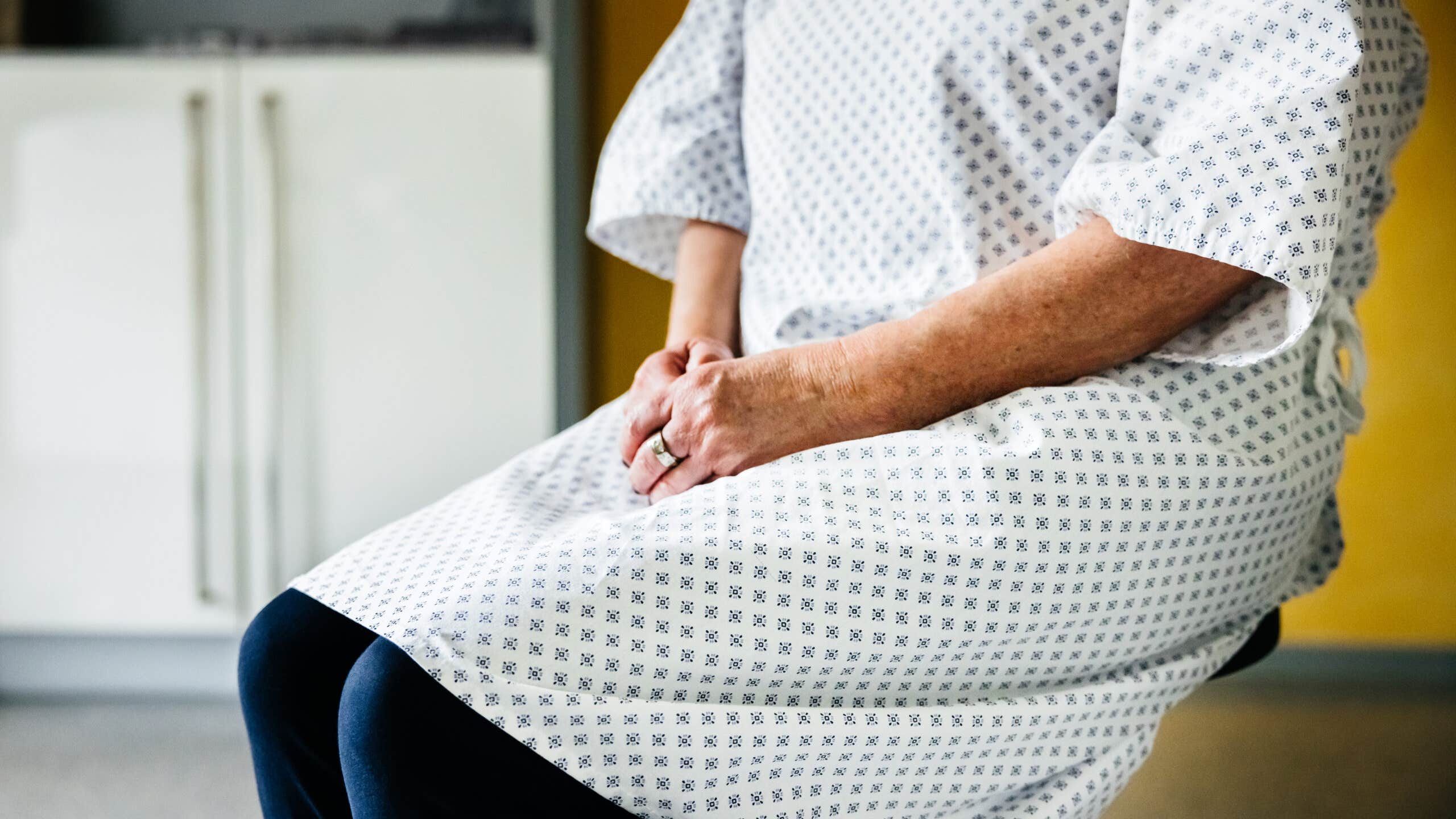 A person waits in a hospital gown with hands folded on their lap.
