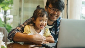 Smiling father holds smiling daughter on his lap while looking at a computer